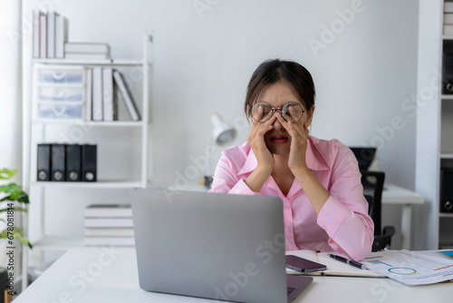 Young Asian female worker working with stress pretends to scratch her head because she is very tired working with laptop and documents on the table.