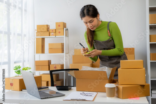 Start a small business SMEYoung Asian woman Business owner using smartphone or laptop receives and checks online orders to prepare boxes of products. Sale.