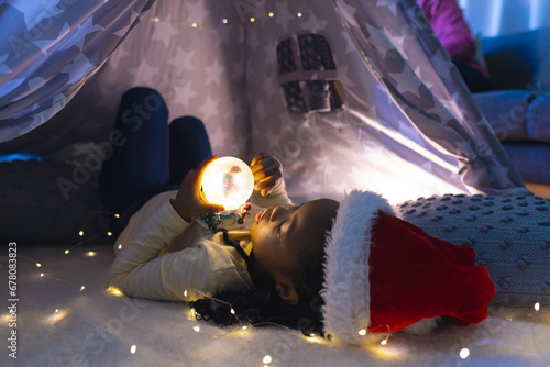 African american girl lying, wearing hat and playing with illuminated christmas snow globe at home photo