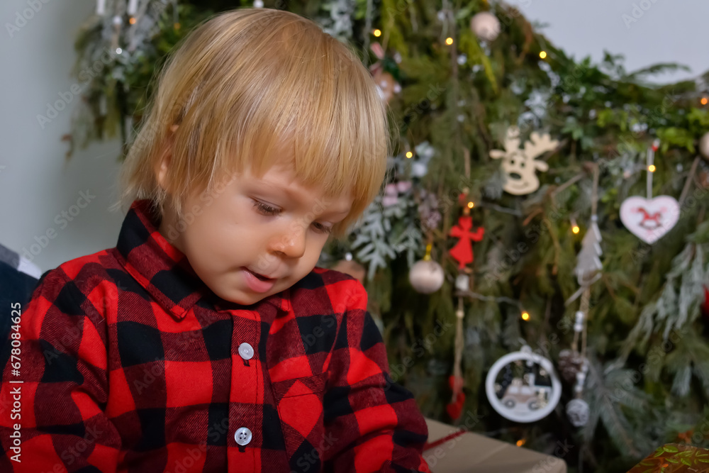 The happy kid is having fun with Christmas presents sitting on the background of the Christmas tree decorated with New Year's garlands. Family Christmas concept The happy kid is having fun with Christmas presents sitting on the background of the Christmas tree decorated with New Year's garlands. Family Christmas concept