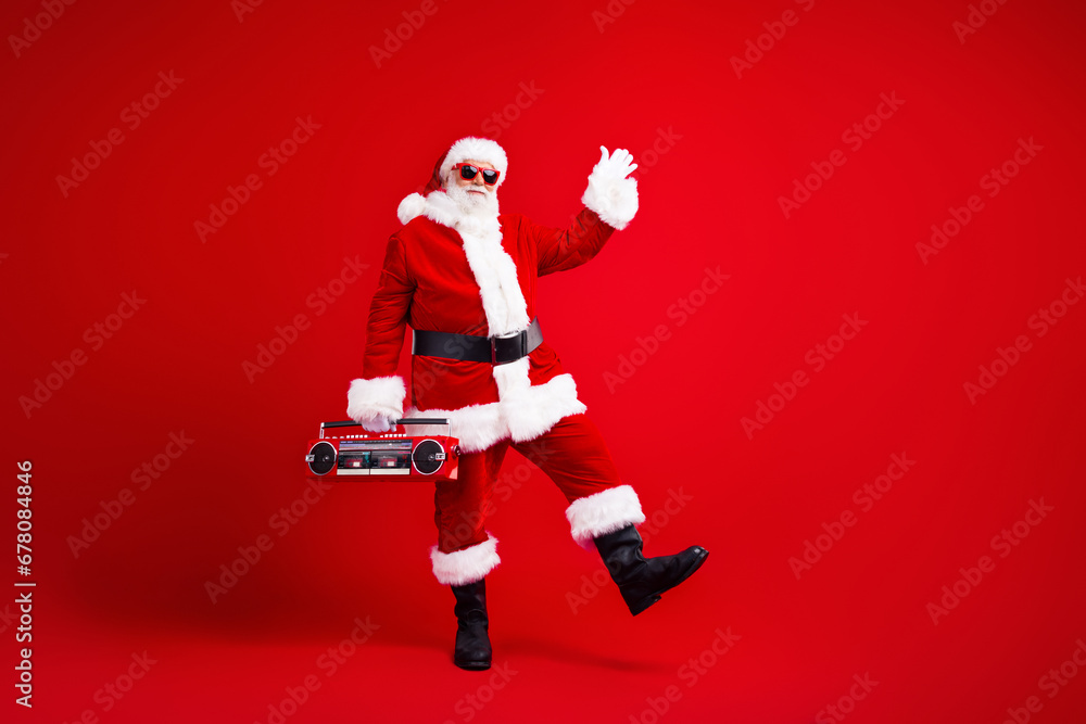 Full length photo of optimistic santa claus in sunglass waving palm say hello hold boombox dancing isolated on red color background Full length photo of optimistic santa claus in sunglass waving palm say hello hold boombox dancing isolated on red color background