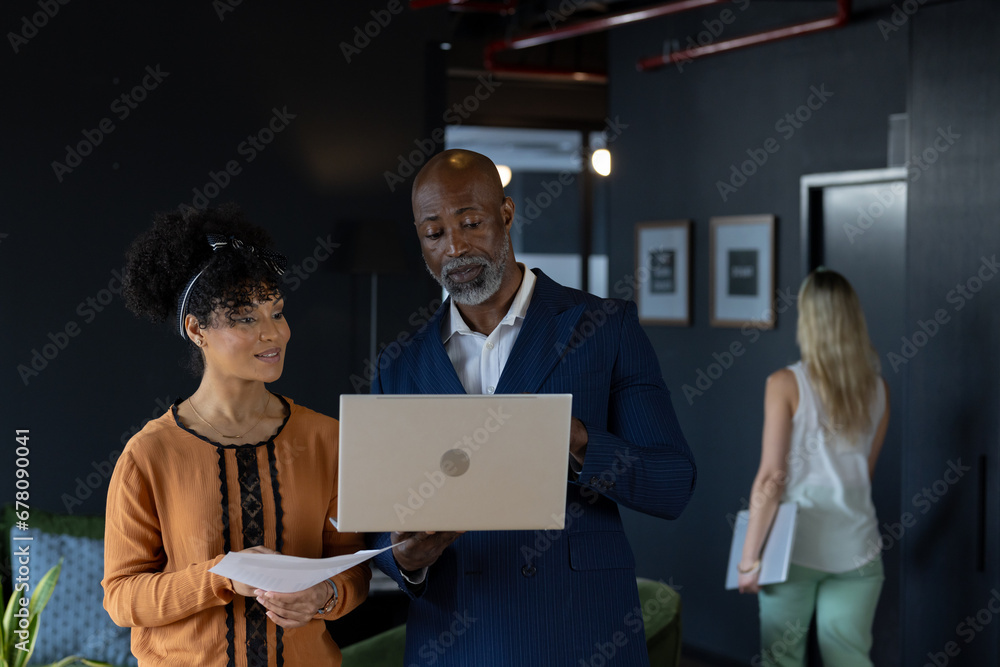 Happy diverse male and female colleague using laptop and discussing document standing in office Happy diverse male and female colleague using laptop and discussing document standing in office