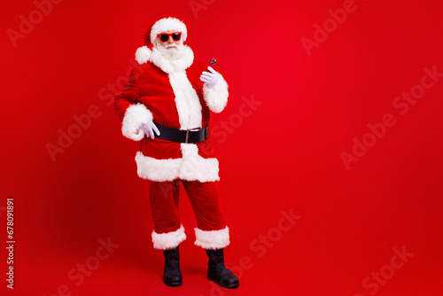 Full size photo of optimistic grandfather wear santa costume in glasses holding smartphone arm on waist isolated on red color background