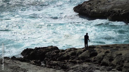 Fisherman Stands On A Rocky Shore And Catches Fish In The Waves