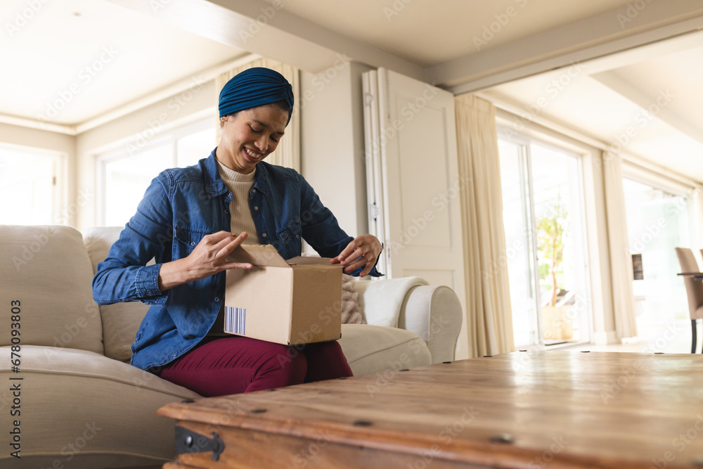 Happy biracial woman in hijab unpacking package in living room Happy biracial woman in hijab unpacking package in living room