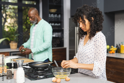Focused mature diverse couple breaking eggs and preparing breakfast in sunny kitchen