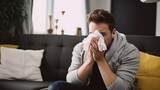 Caucasian man wrapped in warm clothes due to chills has flu sneezing in napkin in modern cozy apartment. Young Caucasian man caught cold and now suffers from flu with runny nose.