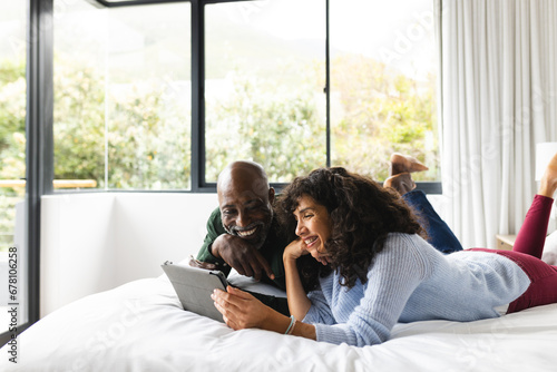 Happy diverse mature couple lying on bed using tablet in sunny bedroom, copy space photo