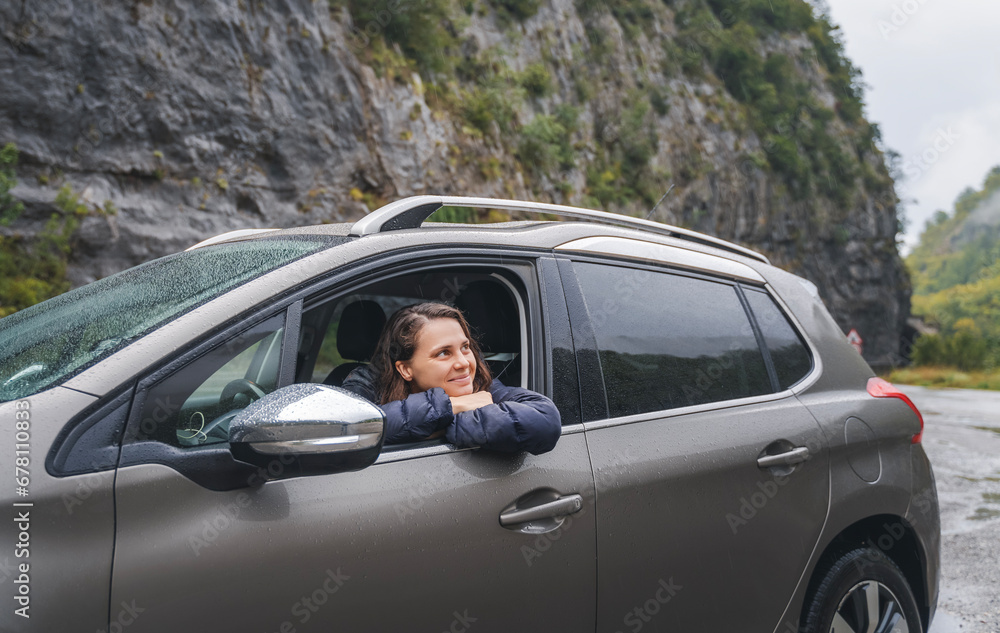 A young smiling woman driver sitting in a car against the backdrop of a mountain in cloudy rainy weather