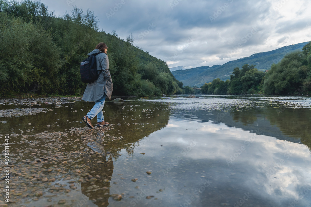 A young woman in a raincoat walking along the bank of a mountain river. Rest and relaxation with nature
