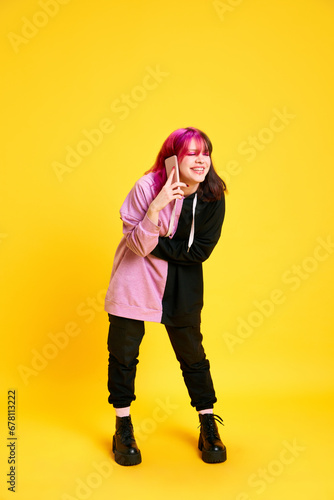 Emotional young girl with pink hair and piercing talking on mobile phone with laugh against vivid yellow studio background. Concept of youth, self-expression, fashion, emotions, communication