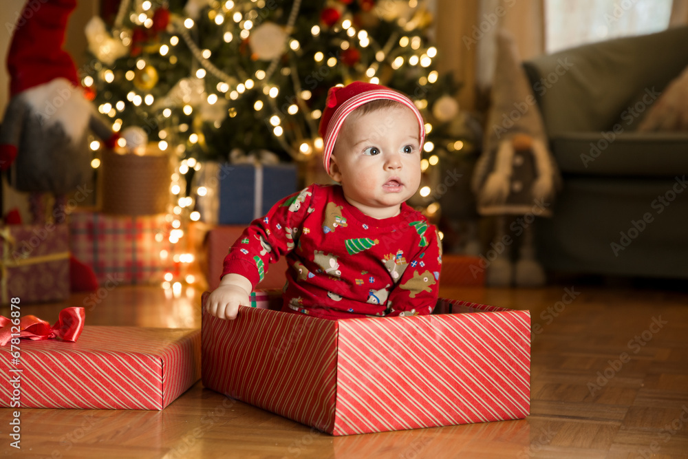 Happy baby boy in a red cap sitting in a gift box on the background of a Christmas tree on Christmas Eve. Happy Holidays, New Year. Cozy warm winter evening at home. Xmas time Happy baby boy in a red cap sitting in a gift box on the background of a Christmas tree on Christmas Eve. Happy Holidays, New Year. Cozy warm winter evening at home. Xmas time