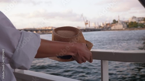 Young woman's hands holding hat with large flat on background of city and Bosphorus in Istanbul in Turkey. She sails on tourist liner or pleasure boat on sea. Tourist shot on deck. photo