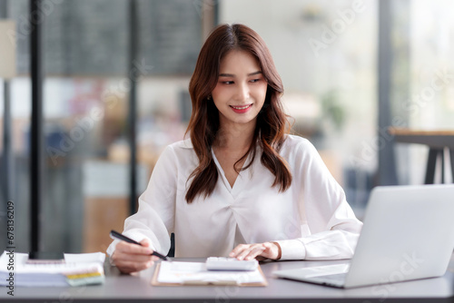 Young Asian businesswoman using laptop computer, taking notes, working project in modern office. Financier working with documents at workplace.