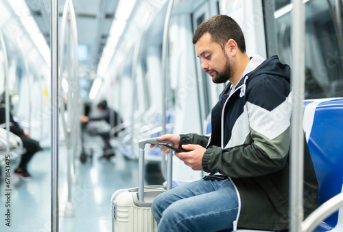 Focused young man traveling with suitcase in empty subway car, sitting on seat and browsing messages on his phone