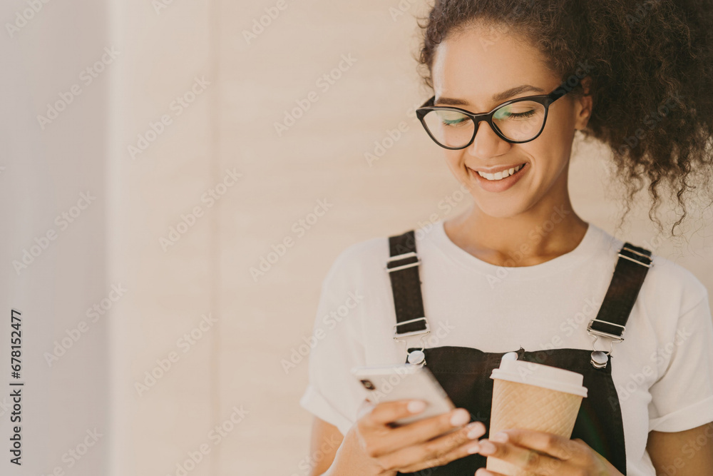 Content young lady with curly hair, glasses, texting on smartphone and holding a coffee Content young lady with curly hair, glasses, texting on smartphone and holding a coffee