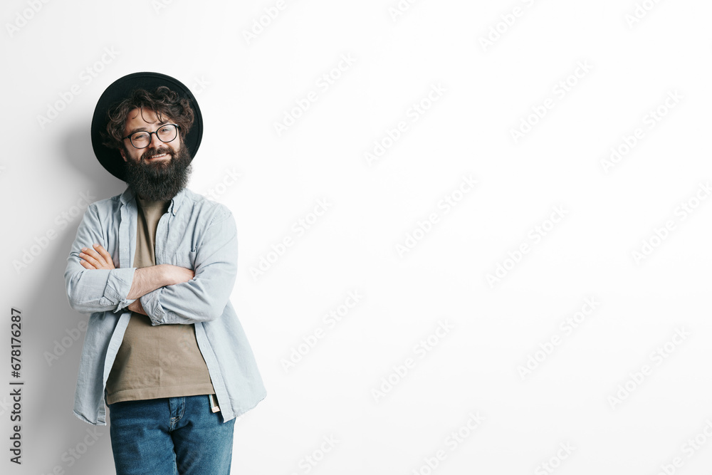 Fototapeta premium Confident bearded man with glasses smiling and standing arms crossed, wearing a stylish hat and casual shirt, against a white background