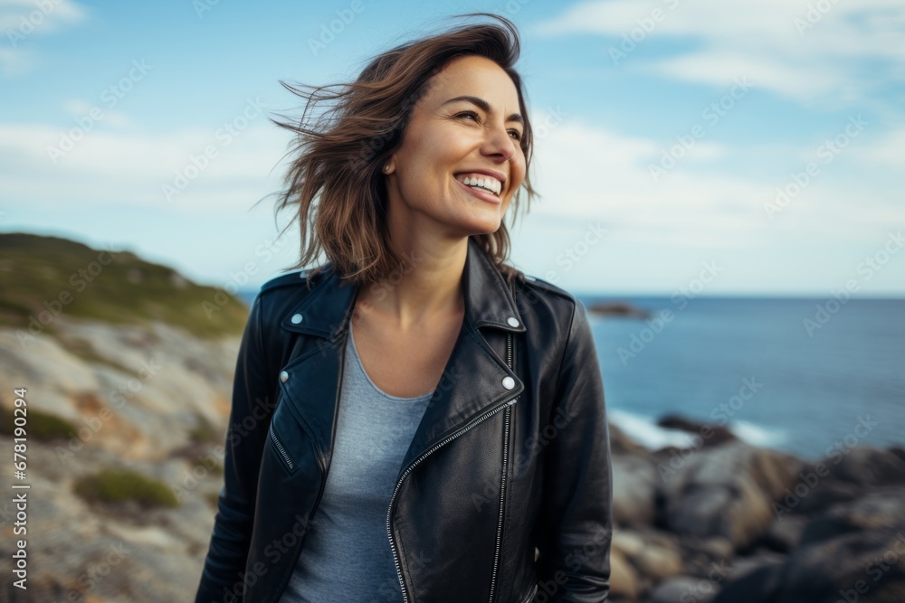 Portrait of a joyful woman in her 40s sporting a classic leather jacket against a serene seaside background. AI Generation