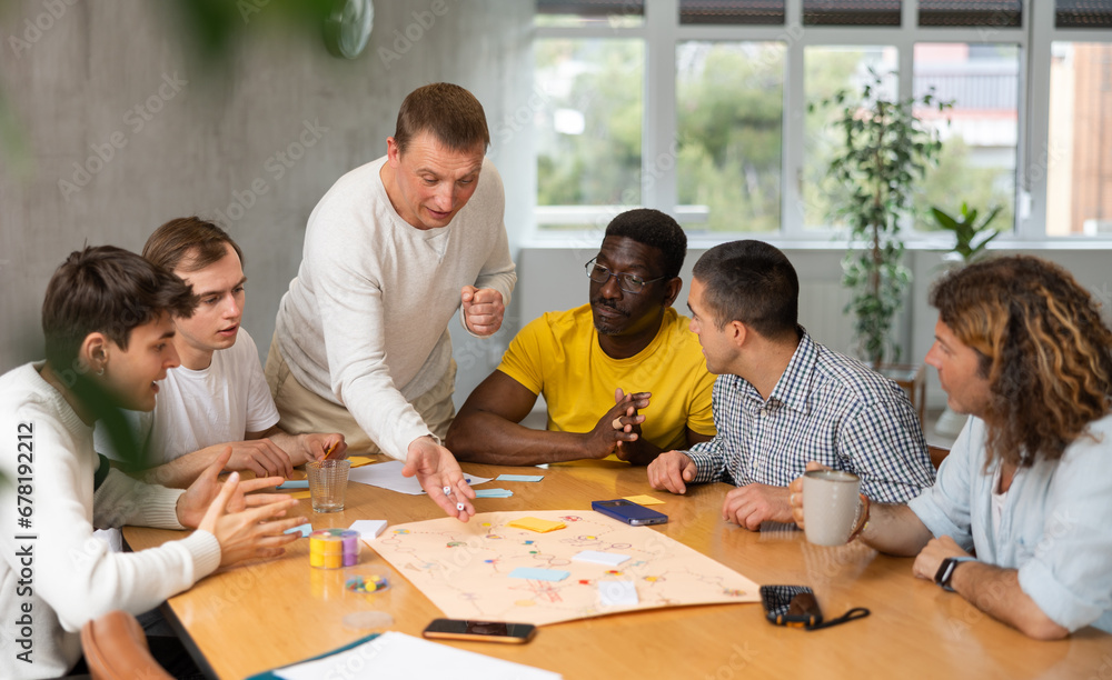 Fototapeta premium Excited man standing on foot playing board game with interested guys sitting around the table