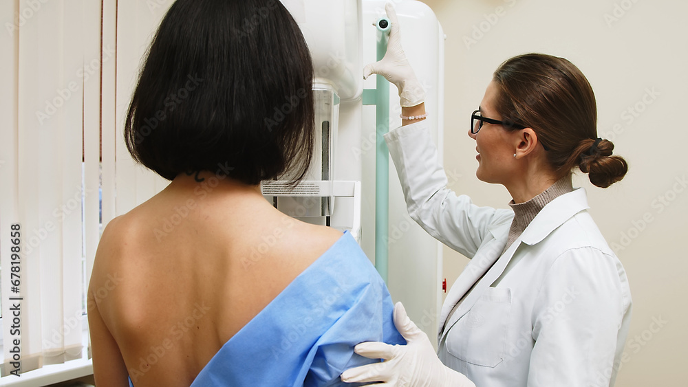 A female doctor in a modern clinic explains the mammography procedure to a topless adult patient undergoing a mammogram. A healthy woman is engaged in cancer prevention in a hospital ward. - obrazy, fototapety, plakaty 