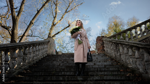 A business woman stands on an ancient staircase, holding white flowers in her hands. Stylish woman in a coat in autumn with a bouquet of white roses. © Roman