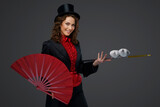 Joyful female illusionist performing magic tricks with white doves while holding a large red fan against a gray background.