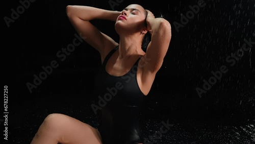 Elegant brunette woman dancing sitting in water and posing under rain at night. Seductive female model, gymnast dancer wearing black body swimsuit, falling raindrops splashes, black background photo