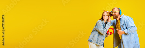 Banner. Father and daughter, parent and kid dressed denim outfit and have fun listening music from one headphones over yellow background.