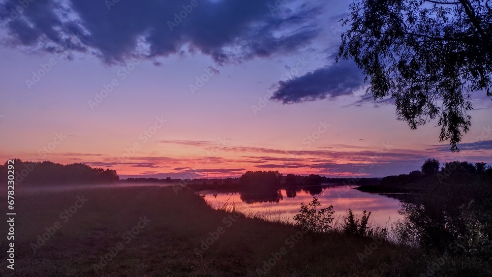 Summer evening, the sun has hidden behind the horizon. The river water reflects a colorful cloudy sky of pink hue and trees. A forest grows on the opposite bank. A fog drifts over the meadow
