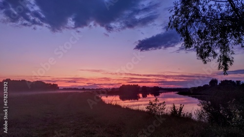 Summer evening, the sun has hidden behind the horizon. The river water reflects a colorful cloudy sky of pink hue and trees. A forest grows on the opposite bank. A fog drifts over the meadow 