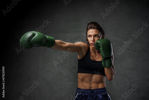 Fit woman boxer in athletic wear, displaying her punch techniques with gloves on, set against a moody background © Fxquadro