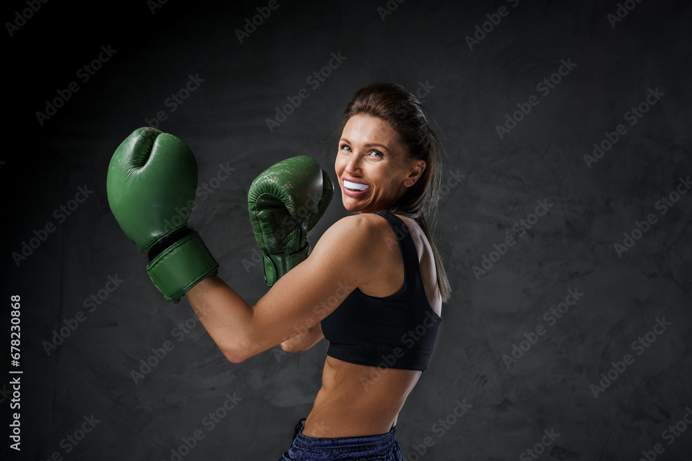 Athletic female fighter with a mouthguard, in sports bra, shorts, and boxing gloves, demonstrates striking skills on a dark background