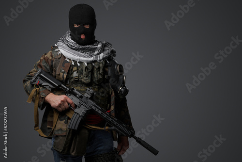 Portrait of a militant from the Middle East, wearing a black balaclava and camo field attire, holding a rifle, set against a gray backdrop