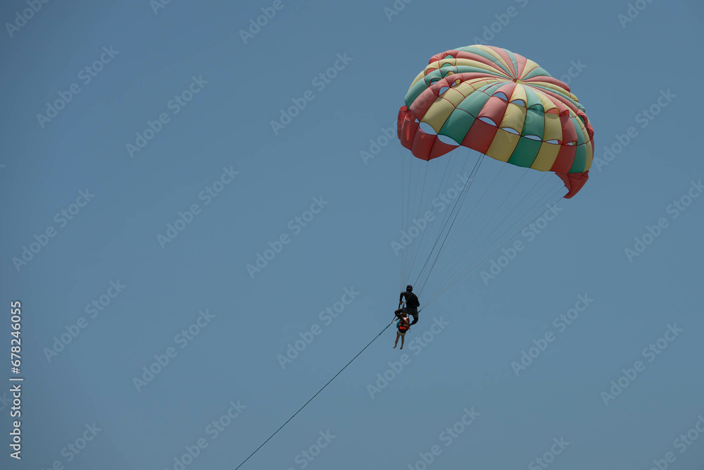 Two persons - man and child are parasailing. Colorful parachute, man holds from behind a child. Close up photo on clear blue sky Two persons - man and child are parasailing. Colorful parachute, man holds from behind a child. Close up photo on clear blue sky
