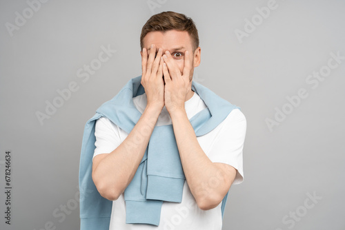 Man looks through fingers. Frightened middle aged guy covers face with hands, palms, looks at camera with scared look. Unpleasant, terrifying creepy scary sight. Isolated on gray, studio portrait.