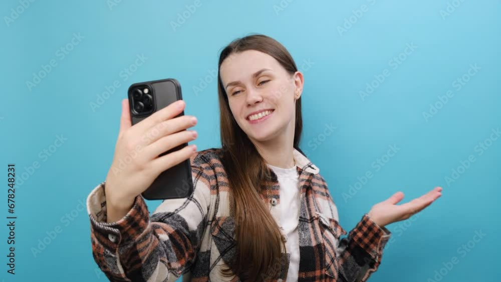 Smiling cute young woman 25s wearing shirt get video call hold use mobile cell phone talk conducting pleasant conversation greet with hand, posing isolated over blue color background wall in studio Smiling cute young woman 25s wearing shirt get video call hold use mobile cell phone talk conducting pleasant conversation greet with hand, posing isolated over blue color background wall in studio