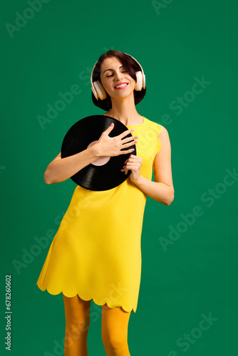 Portrait of young attractive woman dressed style yellow outfit dancing with retro plate while listening music in headphones isolated green background.