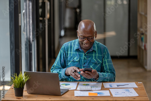 A black senior man in casual clothes uses a cell phone to find information or talk on a touch screen while working at a desk.