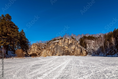 Winter landscape with a large stone on the bank of a frozen river , forest and sky