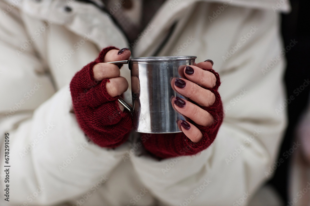Focus on hands of young stylish woman in white winter coat and crimson mittens holding metallic mug with hot tea
