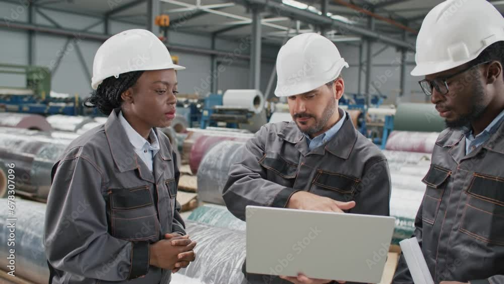 Three mixed-raced workers in helmets and uniforms talking about work while standing in warehouse where stored industrial products. Intelligent foreman holding laptop while speaking with his coworkers. Three mixed-raced workers in helmets and uniforms talking about work while standing in warehouse where stored industrial products. Intelligent foreman holding laptop while speaking with his coworkers.