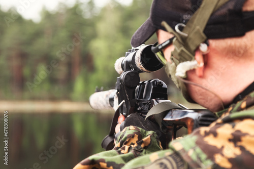 Soldier man sniper in military camouflage uniform shoots from weapon at forest with lake nature backdrop, rear view. Male border guard in country border holding weapon on war. Copy ad text space
