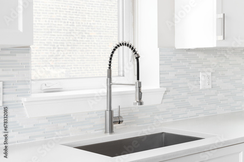 A kitchen faucet detail with a polished stainless steel faucet, a glass and stone mosaic tile backsplash, and white cabinets. photo