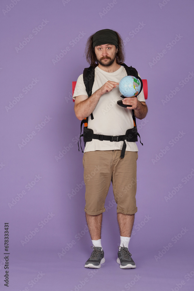 Man tourist in casual clothing with backpack holds globe pointing, looking at camera. Isolated on purple background. Portrait of male traveler. Man tourist in casual clothing with backpack holds globe pointing, looking at camera. Isolated on purple background. Portrait of male traveler.