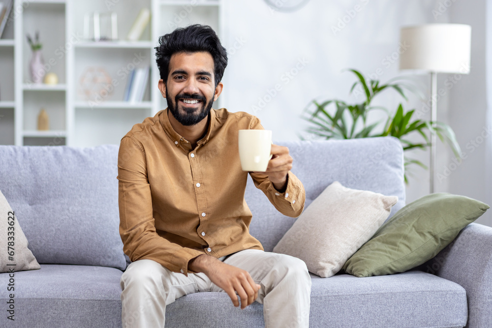 Obraz premium Happy sincere Arab man sitting on sofa at home holding cup in hands, drinking tea or water, concept of healthy lifestyle, healthy habits, rest after working day, smiling looking at camera.