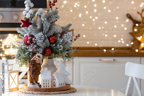 A spruce bouquet with red berries, toys, cotton on a white kitchen table against a beautiful garland. Christmas decorations in the interior. The concept of preparation for the New Year holidays 2024.