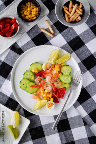 salad with shrimps, cucumbers, egg, red pepper, microgreens and a piece of lime on a white ceramic plate, background - textile in the form of a checkerboard, plates with peppers, grisini sticks nearby photo