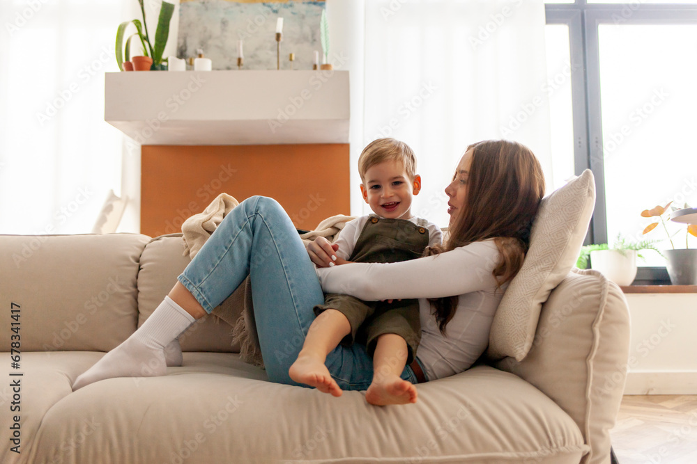 mother with her son sits on the sofa and smiles, woman hugs her child at home, 2 year old boy rests with his parent mother with her son sits on the sofa and smiles, woman hugs her child at home, 2 year old boy rests with his parent