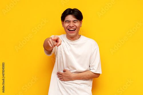 young asian guy in a white t-shirt laughs and points his hand forward on a yellow isolated background photo