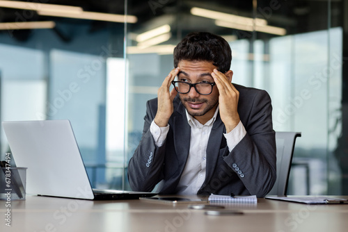 Arab businessman in glasses suffering from headache, sitting in new office working, holding his head, writhing in pain, overtired.